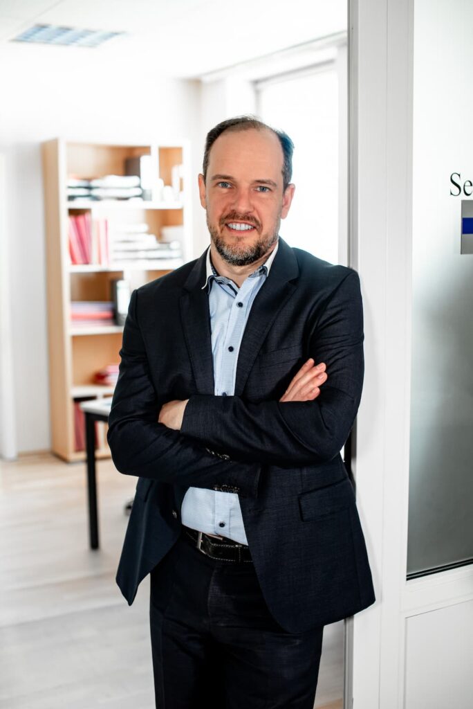Rechtsanwalt Christian Schiebel steht mit verschränkten Armen vor einem Büro und lächelt in die Kamera.